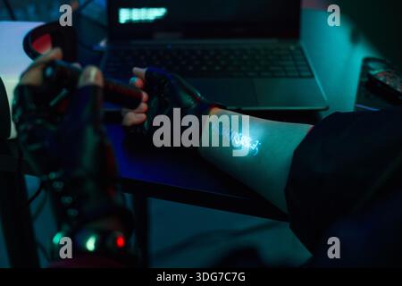 Young adult wearing black gloves holding flashlight projecting hacking code onto forearm while sitting at desk with open laptop, engaging in cyber security or hacker activity Stock Photo