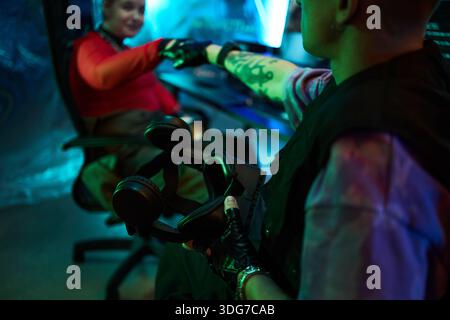 Two young hackers or gamers sitting at computer desk, fist bumping while wearing black gloves, one holding headphones, illuminated by blue and green light in cyber setting Stock Photo