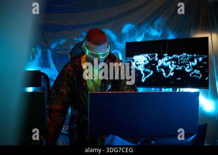 Young adult Caucasian man wearing protective glasses operating multiple computer monitors in dark room, with digital world map on screen, suggesting cybersecurity or hacking activity Stock Photo
