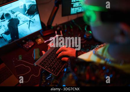 Young adult Caucasian man wearing futuristic glasses sitting at desk typing on wireless keyboard while looking at computer monitor displaying surveillance footage and code Stock Photo
