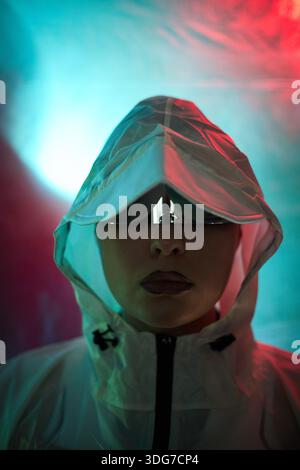 Portrait of young adult Caucasian woman wearing reflective sunglasses and hooded jacket staring directly at camera, illuminated by blue and red lights, evoking hacker theme Stock Photo