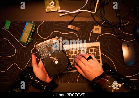 Anonymous young man holding open hard drive while typing on keyboard with other hand, engaging in hacking activity at desk with electronic components and computer mouse visible Stock Photo