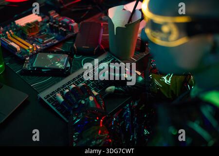 Young man wearing futuristic visor and gloves typing on keyboard surrounded by computer hardware and electronic components, engaging in hacking activity, in dark room Stock Photo