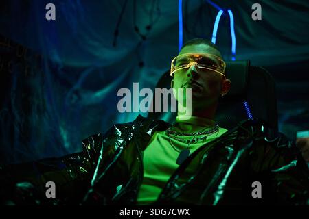 Portrait of young adult Caucasian man wearing futuristic visor sitting in dark room illuminated by neon lights, appearing focused and confident, embodying hacker concept Stock Photo