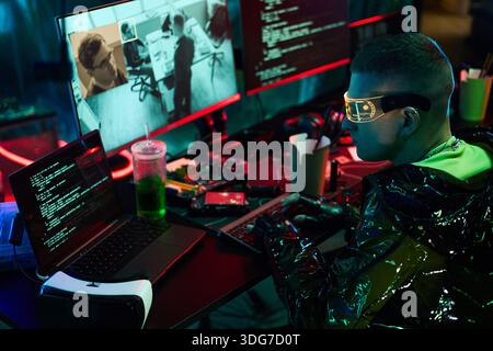 Young adult Caucasian man wearing futuristic glasses sitting at cluttered desk typing on keyboard, multiple monitors displaying code and surveillance footage, engaging in hacking activity Stock Photo
