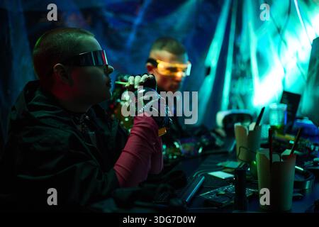 Two young adult Caucasian men, wearing futuristic glasses, sitting at workstation using electronic devices and drinking from metallic bottle, surrounded by computer equipment and cables Stock Photo