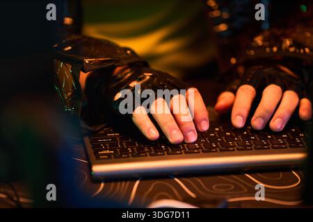 Hands of young man wearing fingerless gloves typing on computer keyboard, hands close up, engaging in hacking activity, illuminated by colorful light, cyber security concept Stock Photo