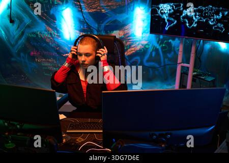 Young adult Caucasian woman sitting at workstation wearing headphones, concentrating on multiple computer monitors with digital world map and neon graffiti background, engaging in hacking activity Stock Photo