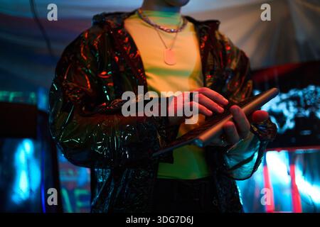 Young adult Caucasian man wearing shiny jacket holding keyboard or using digital tablet in dark room with neon lights, focusing on touchscreen while standing in front of illuminated cyber background Stock Photo