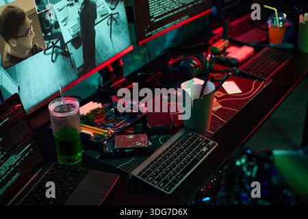Closeup of hacker workspace showing multiple monitors with code and surveillance footage, open takeout container, green drink, computer hardware, keyboard, and headphones scattered on desk Stock Photo