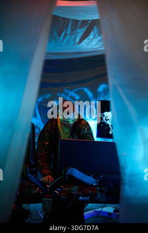 Young Caucasian man wearing neon augmented reality glasses working on multiple computer monitors in dark room neon blue lighting, engaging in hacking activity surrounded by digital equipment Stock Photo
