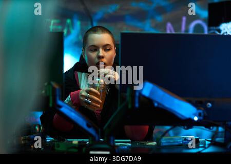 Young adult Caucasian woman sitting at workstation using desktop computers, holding plastic cup with straw, concentrating on screens in dark room with neon graffiti background Stock Photo