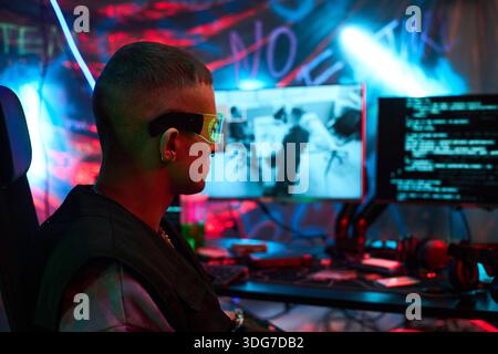 Young adult Caucasian man wearing futuristic glasses, sitting at computer desk, operating multiple monitors with code and surveillance footage, in dark room decorated with graffiti Stock Photo