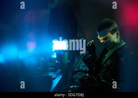 Young adult Caucasian man wearing futuristic visor, sitting at computer desk, holding cable, appearing to hack or engage in cyber crime activity in dark neon lit room Stock Photo