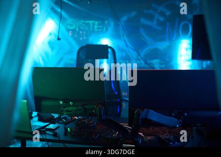 Empty dark room featuring two computer monitors and electronic devices on desk, illuminated by blue neon lights with graffiti on wall, representing hacker workspace environment Stock Photo