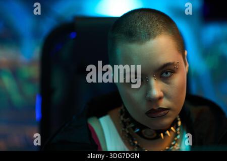 Portrait of young Caucasian woman with shaved head, white eyes and facial piercings sitting in front of computer monitor, wearing spiked choker, looking directly at camera Stock Photo