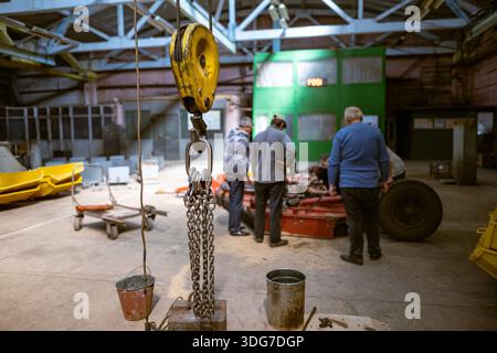 Yellow crane hook and chains in foreground with workers repairing machinery on background. Professional industrial facility interior and engineering m Stock Photo