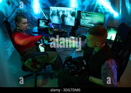 Two young Caucasian woman and man sitting in dark room fist bumping while surrounded by multiple computer monitors displaying code and surveillance footage, engaging in hacking activity Stock Photo