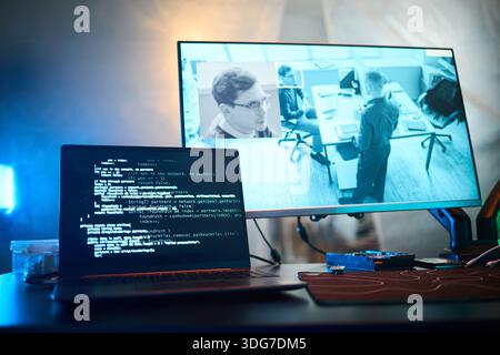 Laptop screen displaying programming code in foreground, large monitor in background showing surveillance footage of office with Caucasian young man and coworkers working at office desks Stock Photo