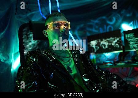 Portrait of young adult Caucasian man, wearing futuristic visor, sitting in dark room illuminated by neon lights, operating multiple computer monitors with digital world map displayed Stock Photo