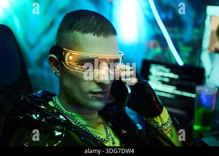 Young adult Caucasian man wearing futuristic glasses sitting at computer desk talking on smartphone surrounded by digital screens with code displaying hacker activity Stock Photo