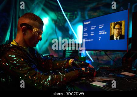 Young adult Caucasian man wearing futuristic visor sitting at desk operating electronic device with circuit boards, hacking computer system, target digital profile displayed on monitor Stock Photo
