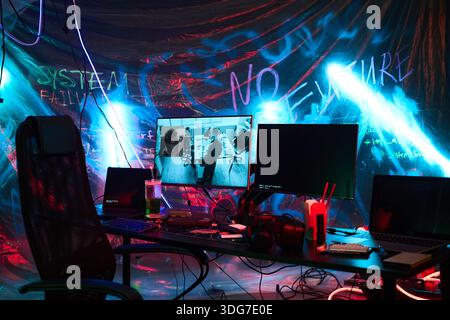 Empty workstation featuring multiple monitors, laptops, headphones, and neon lighting, suggesting hacker environment with digital code and cyber security themes in background Stock Photo