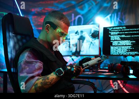 Young adult Caucasian man, wearing protective glasses, sitting at desk, examining computer hardware component in dark room, with multiple monitors displaying code and cyber security data Stock Photo