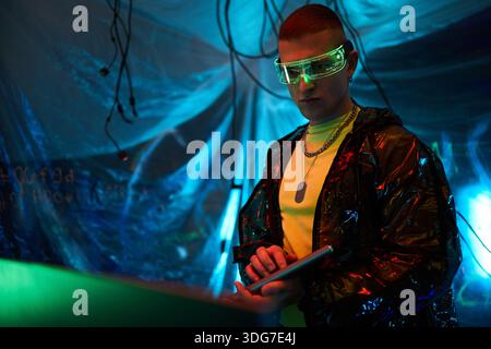 Young adult Caucasian man wearing futuristic glasses standing in dark room with blue lighting operating digital tablet surrounded by hanging cables representing hacker concept Stock Photo
