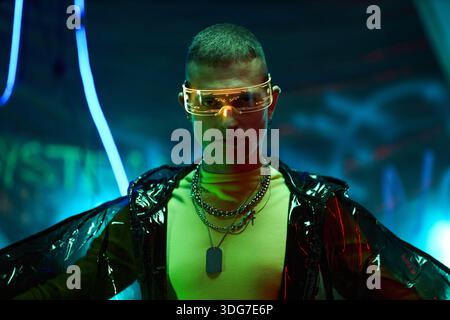 Portrait of Caucasian young adult man wearing futuristic glasses and layered chains standing in neon-lit environment, looking directly at camera with confident expression, hacker concept Stock Photo