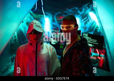 Young adult Asian woman and young adult Caucasian man standing in front of computer monitors displaying code, both wearing futuristic glasses and hooded jackets in dark room Stock Photo