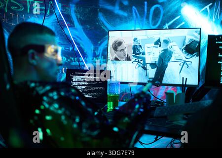 Young adult Caucasian man wearing futuristic glasses sitting at workstation hacking computer system, holding smartphone, monitoring surveillance footage on multiple screens in dark room Stock Photo
