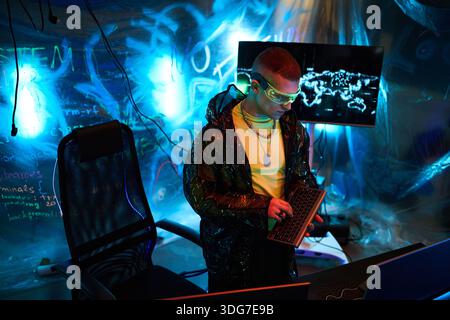 Young adult Caucasian man, wearing futuristic glasses, standing in dark room with neon lights, holding wireless keyboard and looking at computer monitor displaying digital world map Stock Photo