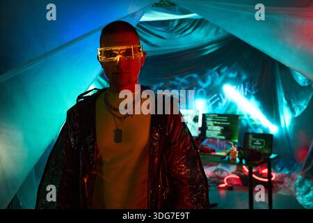 Young adult Black man wearing futuristic glasses standing in front of computer monitors displaying code in dark room with neon lighting, embodying hacker concept, not looking at camera Stock Photo