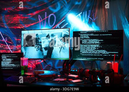 Multiple computer monitors displaying surveillance footage and lines of code on cluttered desk, showing hacker workspace with visible keyboard, headphones, and illuminated background Stock Photo