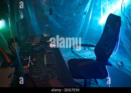 Empty ergonomic chair positioned at workstation with dual monitors displaying computer hacking code, multiple headphones and electronic devices scattered on desk, illuminated by blue neon lighting Stock Photo