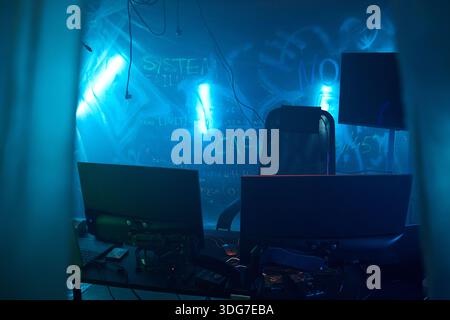 Empty office chair surrounded by multiple computer monitors and laptops, in dark room with blue neon lights and graffiti on wall, suggesting hacker workspace or cybercrime scene Stock Photo