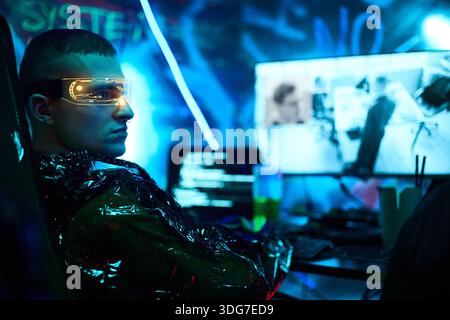 Young adult Caucasian man wearing futuristic visor sitting at computer desk hacking system, illuminated by neon lights, multiple monitors displaying code and surveillance footage Stock Photo