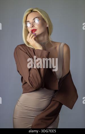Thoughtful studio portrait of blonde woman touching lips, wearing glasses, blazer and beige dress. Minimal gray background with copy space. Stock Photo