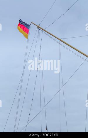German national flag flutters in strong wind on high mast of historic sailing vessel with complex rigging, steel wires and yards against overcast clou Stock Photo