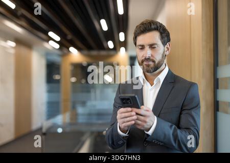 Businessman in a suit standing in an office corridor, focused on typing and checking messages on his smartphone, connecting, networking and managing work digitally while on the move Stock Photo