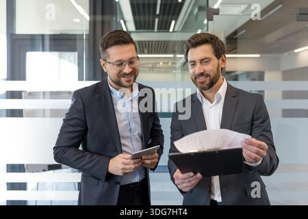 Two smiling professional businessmen standing in a modern office, collaborating and reviewing documents on a clipboard while one holds a tablet, focusing on partnership and planning Stock Photo