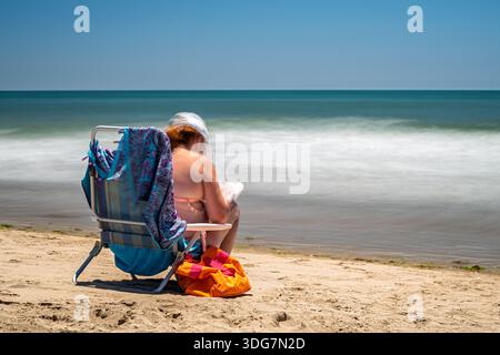 A woman relaxes, solving a crossword puzzle while sitting in a beach chair on Isla Canela, Ayamonte, Huelva, Spain during a sunny day. Long exposure. Stock Photo