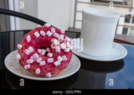 Pink glazed donut with marshmallows on plate beside white coffee cup Stock Photo