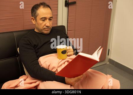 Man reading a book while resting at home with a blanket and a mug Stock Photo