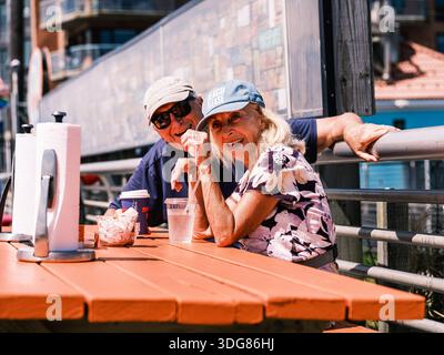 Elderly couple relax at an outdoor table on the Long Beach boardwalk, enjoying cold drinks on a sunny summer day in New York. Stock Photo