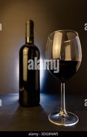 Close-up of a red wine glass in sharp focus with a dark wine bottle blurred behind, moody studio backlight and copy space. Stock Photo