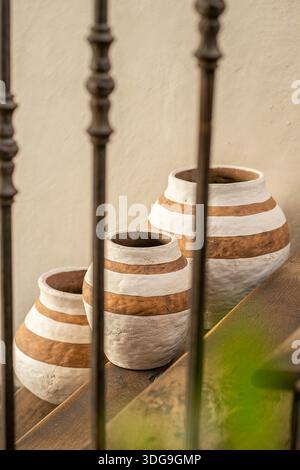 Three handcrafted clay vases with white horizontal stripes arranged on a wooden ledge in a rustic interior setting. Stock Photo