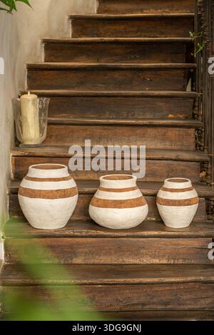 Three handcrafted clay vases with white horizontal stripes arranged on a wooden ledge in a rustic interior setting. Stock Photo