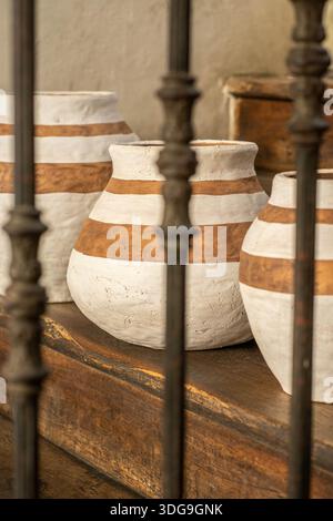Three handcrafted clay vases with white horizontal stripes arranged on a wooden ledge in a rustic interior setting. Stock Photo
